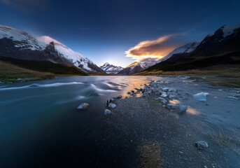 Poster - A river flows through a valley with snow capped mountains.  A beautiful orange cloud covers the sky.