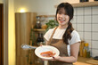 © Prathankarnpap - Beautiful young woman in apron holding a plate with sausages and bacon standing in a kitchen