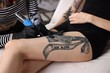 © New Africa - Professional artist tattooing woman's leg with machine in salon, closeup