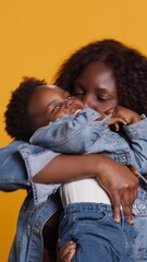 Wall Mural - Vertical Video Smiling african american mom carrying her sweet little boy in studio, embracing him and bonding in front of the camera. Cheerful mother and cute small baby posing together, loving mommy