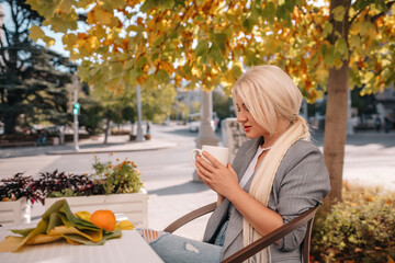 Wall Mural - A blonde woman is sitting at a table with a cup of coffee and a plate of fruit. She is smiling and enjoying her time outside.