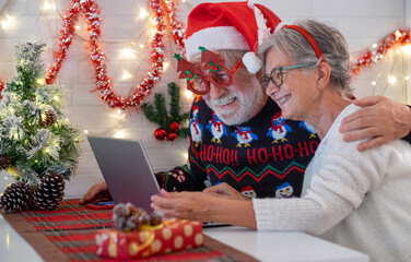 Senior couple in Christmas sweeter and santa hat using laptop computer together. Elderly happy couple celebrate Christmas event with decorated desk and wall. Merry Christmas and happy new year