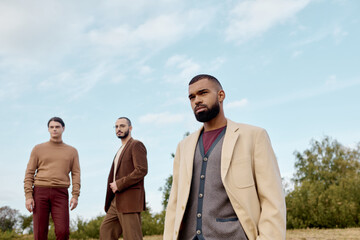 Three handsome men in fashionable autumn attire pose confidently in a tranquil field under clear skies.