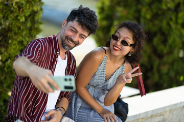 Happy couple taking a cheerful selfie together outdoors with trees in the background, both smiling and making peace signs while enjoying a sunny day.