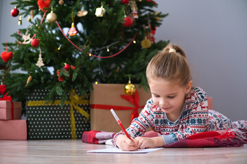 A cute child in Christmas pajamas writes a letter to Santa near the Christmas tree