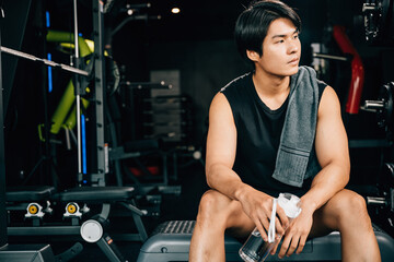 Portrait of Asian young man trained body in black sportswear sitting for rest holding water or protine in fitness gym, Weight training, healthy lifestyle concept