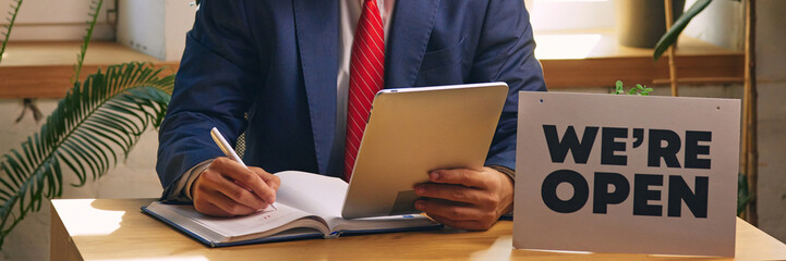 Businessman in formal wear sitting at desk with we are open sign and signing documents, checking information with tablet. Cropped image. Concept of new business, startup, achievement, progress