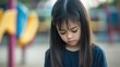 © Lion - Stock minimalist photograph of a sad young Asian girl standing alone in an empty playground, looking down at the ground, with softly blurred equipment in the background