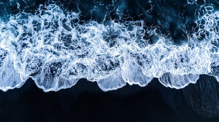 Drone aerial view of the sea or ocean waves splashing on a dark navy blue or gray sand beach with beautiful transparent clear water