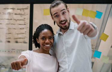 Half length portrait of cheerful multicultural male and female partners standing in office room and showing ok while smiling at camera, positive diverse coworkers enjoying entrepreneurship indoors