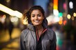 © Markus Schröder - Portrait of a joyful indian woman in her 30s wearing a functional windbreaker while standing against background of bright city lights