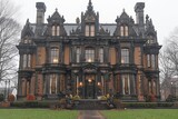 A grand, ornate historical building with detailed Victorian architecture and intricate designs stands surrounded by trees and a well-maintained lawn