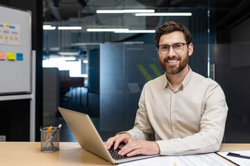 Portrait of a young successful man in glasses and a light shirt sitting in the office at the table, working on a laptop, smiling and looking at the camera