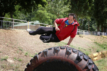 A guy in a red sweater jumps over a large black rubber tire in a parkour style with an acrobatic trick in the stadium against the background of plants and trees in the summer	
