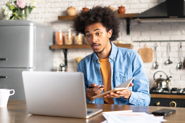 Young confident African American man working from home sitting in the kitchen with his laptop computer, participating in video conference, having online meetup with his coworkers, employee or client