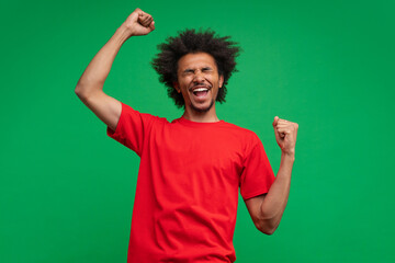 Studio shot of euphoric African American young man screaming yes with triumph, clenching and raising fists up, celebrating victory, isolated on green background