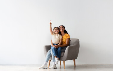 Wall Mural - Check This. Happy Young Mother And Little Daughter Sitting In Armchair And Pointing Up, Cheerful Middle Eastern Mom And Female Child Demonstrating Copy Space Above Their Heads While Resting In Chair