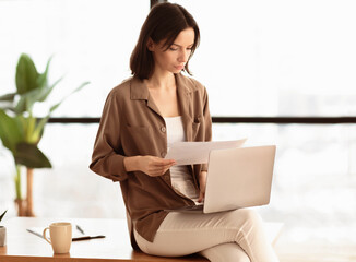 Aufkleber - Pensive young woman sitting on desk at office holding paper documents, working on laptop, typing, copyspace