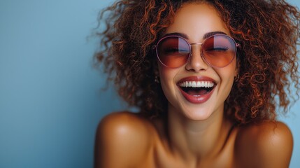 Wall Mural - Young woman with curly hair laughs joyfully while wearing stylish sunglasses against a bright blue background