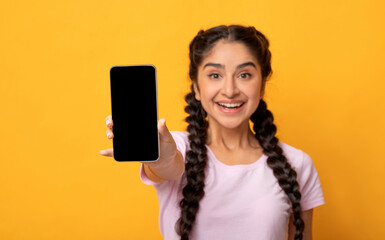 Trendy Mobile Phone. Portrait of cheerful young indian woman holding cellphone with black blank screen in hand, showing device. Gadget with empty free space for mock up, selective focus, blur