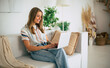 © My Ocean studio - Cute blonde woman sits on a comfortable couch in a modern, stylish living room, working on her laptop. She is smiling, wearing a striped shirt and jeans, surrounded by cozy home decor and plants.
