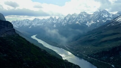 Aufkleber - Sunlight is illuminating the snow capped peaks of a majestic mountain range, casting shadows over a valley where a river meanders through lush greenery