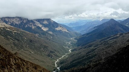 Poster - Winding road is cutting through a valley in the middle of mountains, surrounded by lush green forest, with a cloudy sky