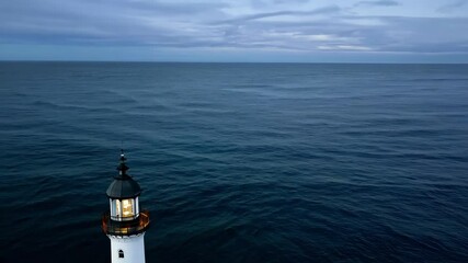 Poster - Lighthouse beaming light at dusk over the ocean, with a tranquil evening sky and crashing waves