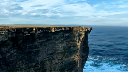 Poster - Sunlight is illuminating the top of a dramatic cliff face jutting out over blue ocean waves, creating a sense of awe and wonder