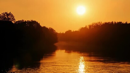 Aufkleber - The setting sun is bathing the landscape in a warm orange light, creating a beautiful contrast with the dark silhouettes of the trees lining the riverbanks