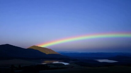 Poster - Colorful rainbow arches over a mountain valley at sunset, painting the sky in vibrant hues. The serene scene radiates peace and beauty in nature