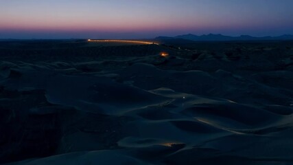 Poster - Long exposure photograph of a winding road illuminating the dramatic landscape of sand dunes at night. The sky is a beautiful gradient of pink and blue, with mountains in the distance