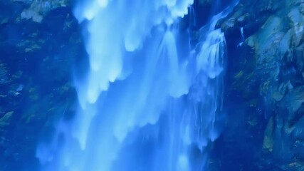 Poster - Water flowing over cliff edge, appearing frozen, gaining momentum in captivating nature photo. Powerful cascade of blue water against rocks and moss creates serene landscape