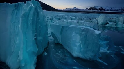 Aufkleber - Large, luminous blue iceberg floats in dark water, reflecting the twilight sky and surrounding mountains. The serene beauty of the scene highlights the fragility of nature