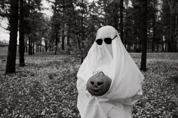 Wall Mural - Woman in white sheet holding jack o'lantern against autumn forest background. Halloween costume. Black and white photo. 