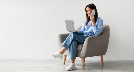 Poster - Full length of Caucasian lady using laptop and speaking on smartphone while sitting in armchair against white studio wall, free space. Lovely young woman having phone conversation, working on pc