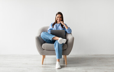 Poster - Millennial Caucasian lady in casual wear using laptop, sitting in armchair, having online conference against white studio wall. Young woman communicating online, taking part in webinar