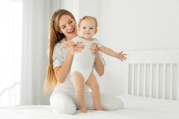 Happy young woman sitting on bed with her little daughter, baby standing with mommy's help, mother looking at child with charming smile