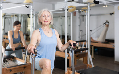 Canvas Print - Elderly active woman doing stretching of body muscles sitting on special chair in pilates room