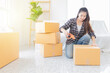 © jokekung - Young woman in plaid shirt seals boxes with tape in bright room. White couch, wooden ladder, and potted plant hint at fresh start in new home. use Adhesive tape packaging paper box. logistic business.