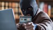© sirisakboakaew - Masked online criminal using a laptop to commit financial fraud, causing disaster