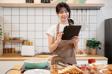 A beautiful Asian woman is looking at a recipe on her digital tablet while cooking in the kitchen.