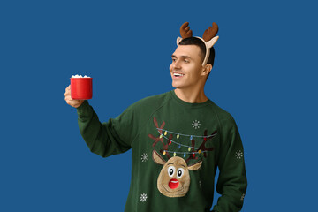 Poster - Young man in reindeer horns holding cup of coffee with marshmallows on blue background