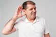 © fotofabrika - Elderly man smiling, playfully holding his hand to his ear while listening intently against a light background