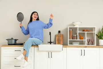 Canvas Print - Happy young woman with frying pan sitting on counter in light kitchen