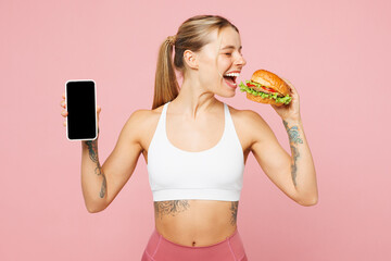 Young woman wear white top casual clothes hold eat bite burger use blank screen mobile cell phone isolated on plain pastel pink background. Proper nutrition healthy fast food unhealthy choice concept.