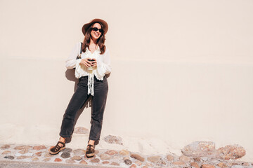 Lifestyle portrait of young stylish woman dressed in white blouse. Model walking with coffee cup in sunny day. Beautiful smiling female in hat. Cheerful and happy. Takeaway coffee, near  wall