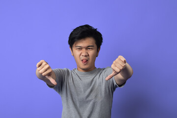 A young Asian man in a gray t-shirt is standing in front of a purple background. He has a neutral expression on his face and is giving two thumbs down with both hands extended forward