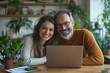 © Danica - Smiling middle-aged business couple using a laptop for a video call in the office, working together at a wooden desk with documents and plants in the background. The smiling mature male manager is tal