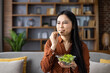 © Liubomir - Asian woman eating salad with dissatisfied expression, contemplating diet. Seated on couch in contemporary interior setting, representing healthy lifestyle choices. Concept of wellness journey.